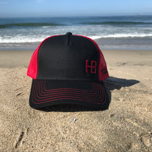 Load image into Gallery viewer, Red embroidered HB in the lower left corner of the front part of this low profile, snapback trucker hat. Huntington Beach California is embroidered on the left side of the hat in black stitching.  5 panel, Low profile, structured firm front panel, pre-curved visor, red mesh back, cotton blend twill front consisting of 65% Polyester and 35% Cotton.